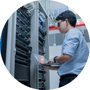 Rechenzentrum_rund Person arbeitet mit einem Laptop vor einem offenen Serverschrank in einem Rechenzentrum. Mehrere Server und Netzwerkgeräte sind in den Racks installiert, Kabel verlaufen geordnet zwischen den Geräten.